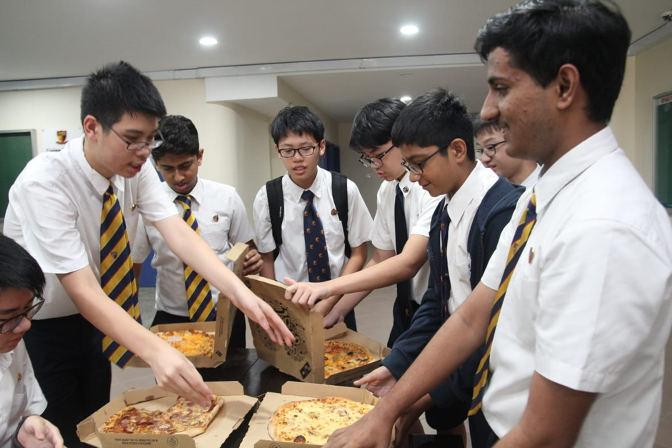 A group of male students in school uniforms crowd around a table, enthusiastically reaching for slices of pizza at what appears to be a celebratory gathering.